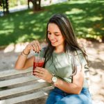 Smiling woman sitting on a bench enjoying a cold beverage in a sunny park.