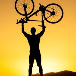 Silhouette of a strong cyclist holding a bike over their head against a stunning sunset.