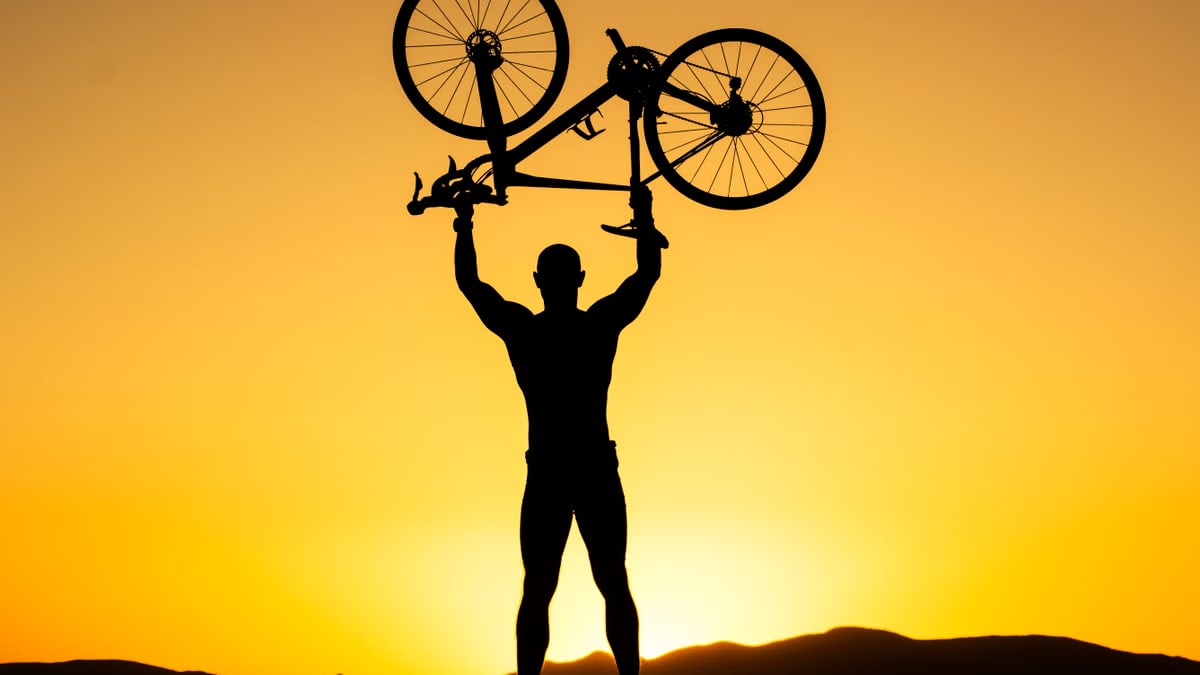 Silhouette of a strong cyclist holding a bike over their head against a stunning sunset.
